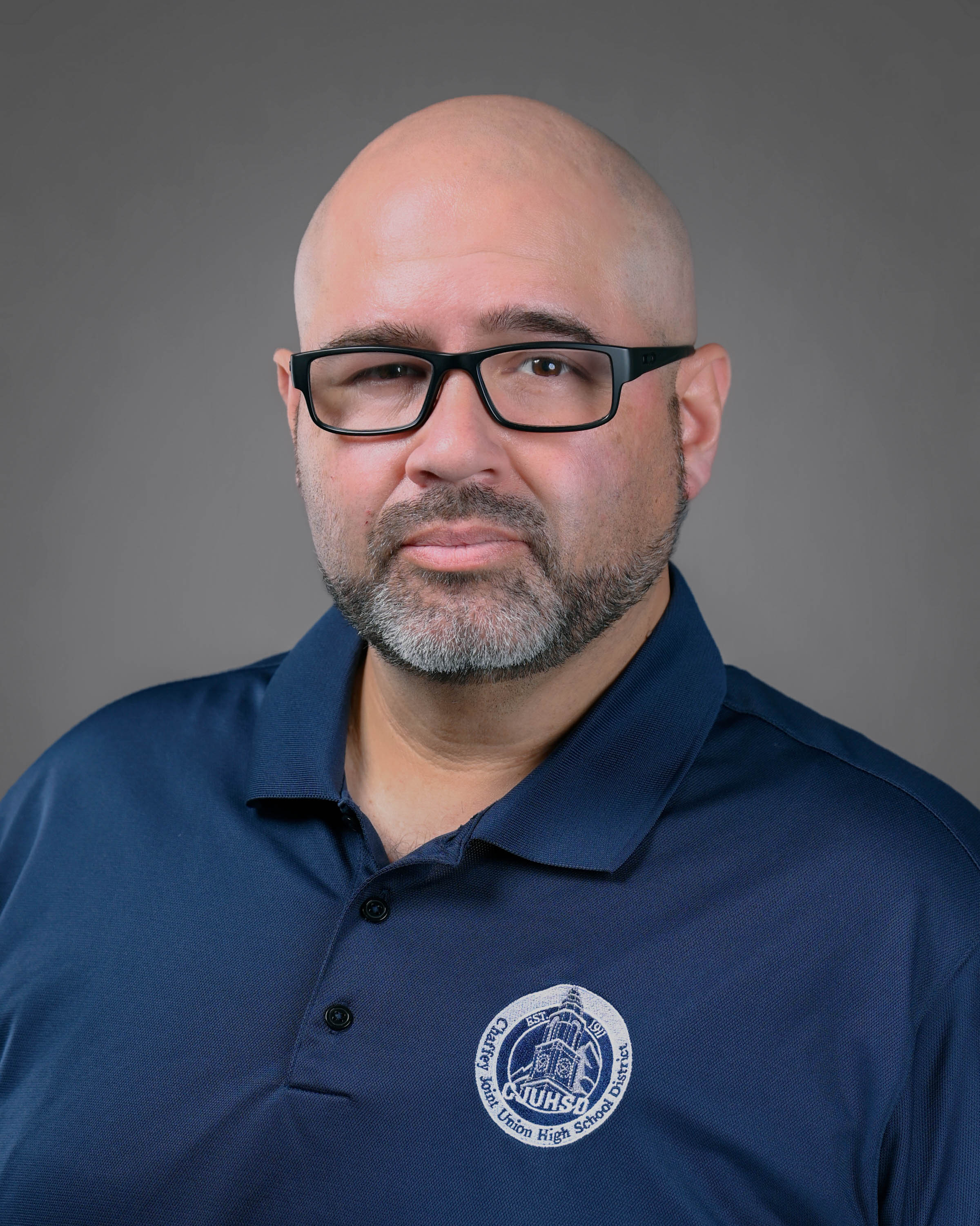 Clean cut man with a short beard wearing eyeglasses with a blue polo shirt on.